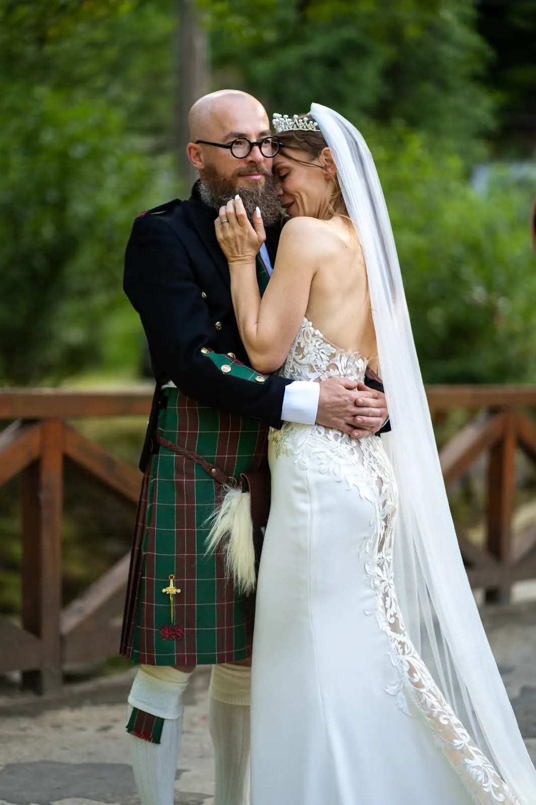 Braut schmiegt sich an ihren Bräutigam im traditionellen schottischen Kilt, im Hintergrund Bäume und ein Holzgeländer